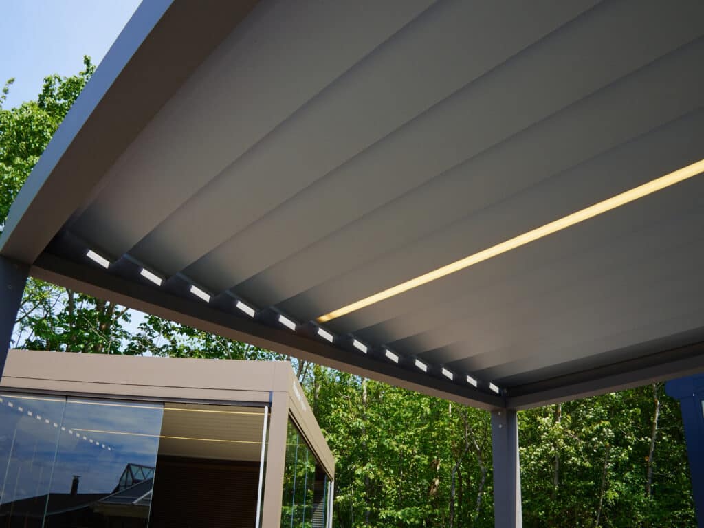 Pergola bioclimatique moderne: Lattes, éclairage LED Pergola bioclimatique grise avec lattes ajustables, bande lumineuse LED jaune et spots encastrés. Arbres verts et ciel bleu en fond.