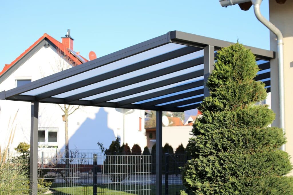 Pergola Moderne Grise : Design, Protection & Toit Translucide Pergola moderne grise à toit translucide devant une maison blanche aux tuiles rouges sous ciel bleu. Jardin avec grand conifère et clôture métallique.
