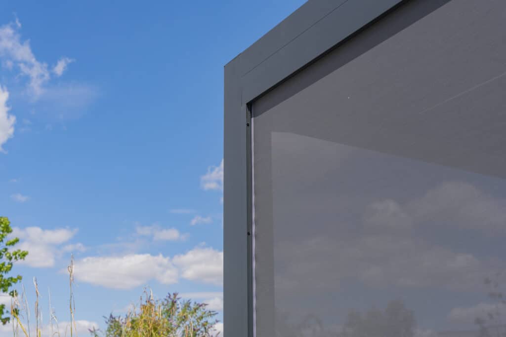 Pergola grise avec stores: Aménagement extérieur moderne Coin de pergola moderne grise avec stores latéraux en tissu sous un ciel bleu nuageux. Végétation verte en arrière-plan.