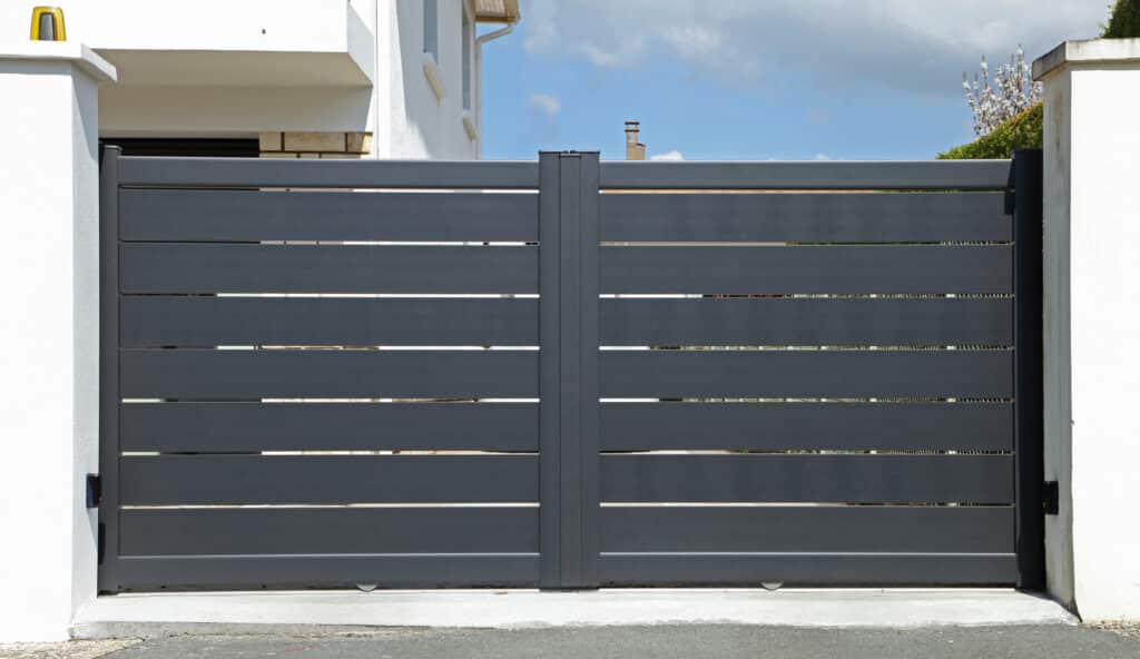 Portail coulissant alu gris anthracite moderne avec sécurité Modern gris anthracite coulissant aluminium gate avec capteurs sécurité, feu clignotant et roues visibles. Maison et ciel bleu en arrière-plan.