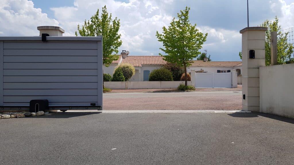 Portail coulissant moderne: Accès rue résidentielle avec maisons Portail coulissant gris ouvert sur rue résidentielle. Maisons aux toits tuilés, arbres verts et ciel nuageux. Le numéro 3 est visible.