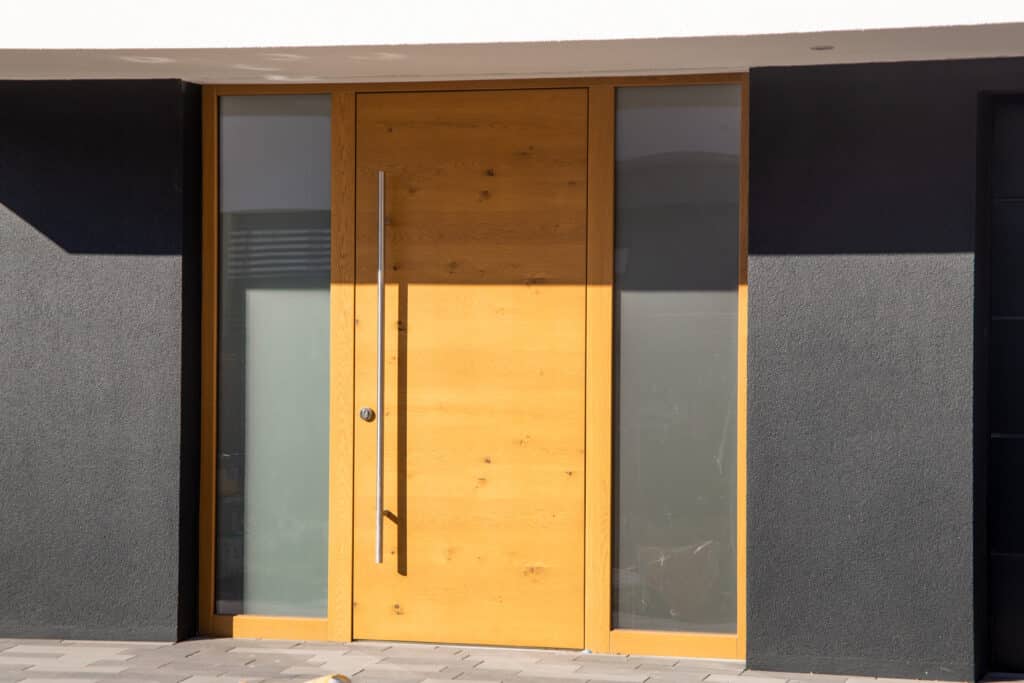 Porte d'entrée moderne en bois : design et élégance Porte d'entrée moderne en bois clair avec poignée argentée et fenêtres latérales. Murs gris foncé, plafond blanc, sol pavé, ensoleillé.