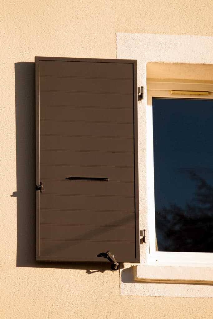 Volet Marron Moderne et Façade Beige Ensoleillée avec Ombre Volet marron fermé sur façade beige texturée, avec poignée et loquet noirs. Lumière du soleil intense, ombres nettes. Fenêtre blanche à côté, reflet d'arbre.