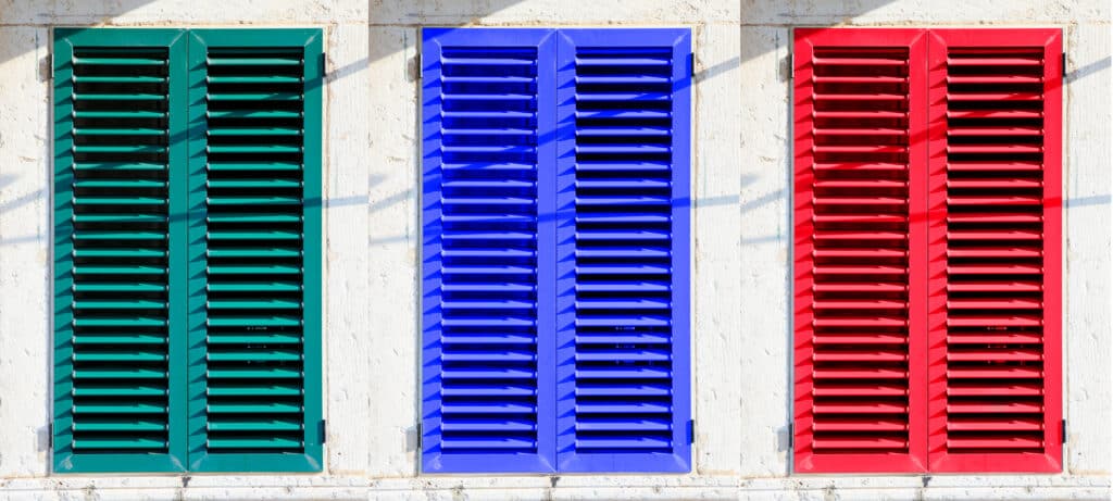 Volets colorés vert, bleu et rouge sur mur blanc Trois volets persiennes extérieurs aux couleurs vives : vert sarcelle, bleu roi et rouge vif, devant un mur blanc texturé en plein soleil.