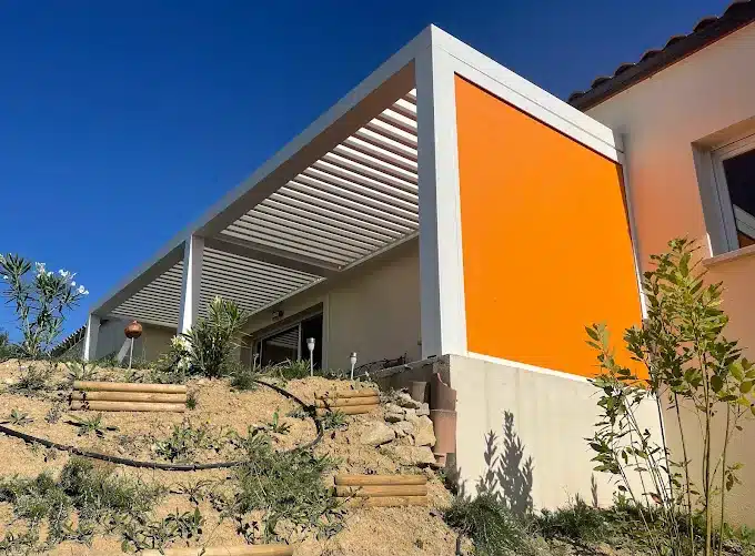 Pergola bioclimatique blanche à lames orientables et store vertical orange devant une maison moderne.